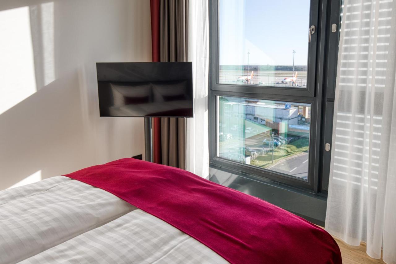 Intercityhotel Berlin Airport Ber Terminal 1 & 2 쇼네펠드 외부 사진 The photo shows a modern hotel room with a large bed covered in white linens and a burgundy throw blanket. In the background, there is a large window that lets in natural light, offering a view of the outside. A flat-screen TV is mounted on the wall 