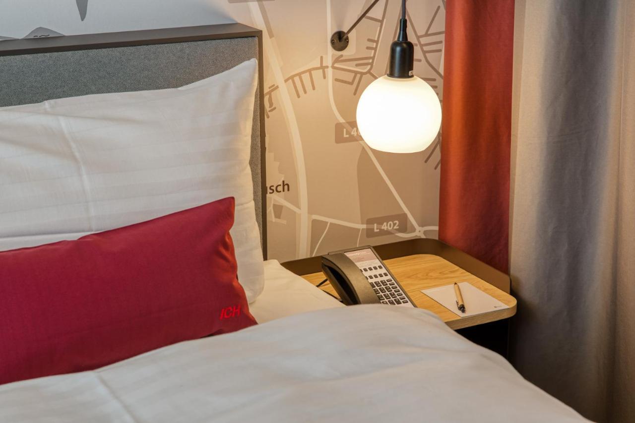 Intercityhotel Berlin Airport Ber Terminal 1 & 2 쇼네펠드 외부 사진 The photo shows a cozy bedroom setup. There's a neatly made bed with white bedding and a red decorative pillow. Beside the bed, a small bedside table holds a telephone and a notebook or notepad, along with a pen. A round pendant light hangs above the