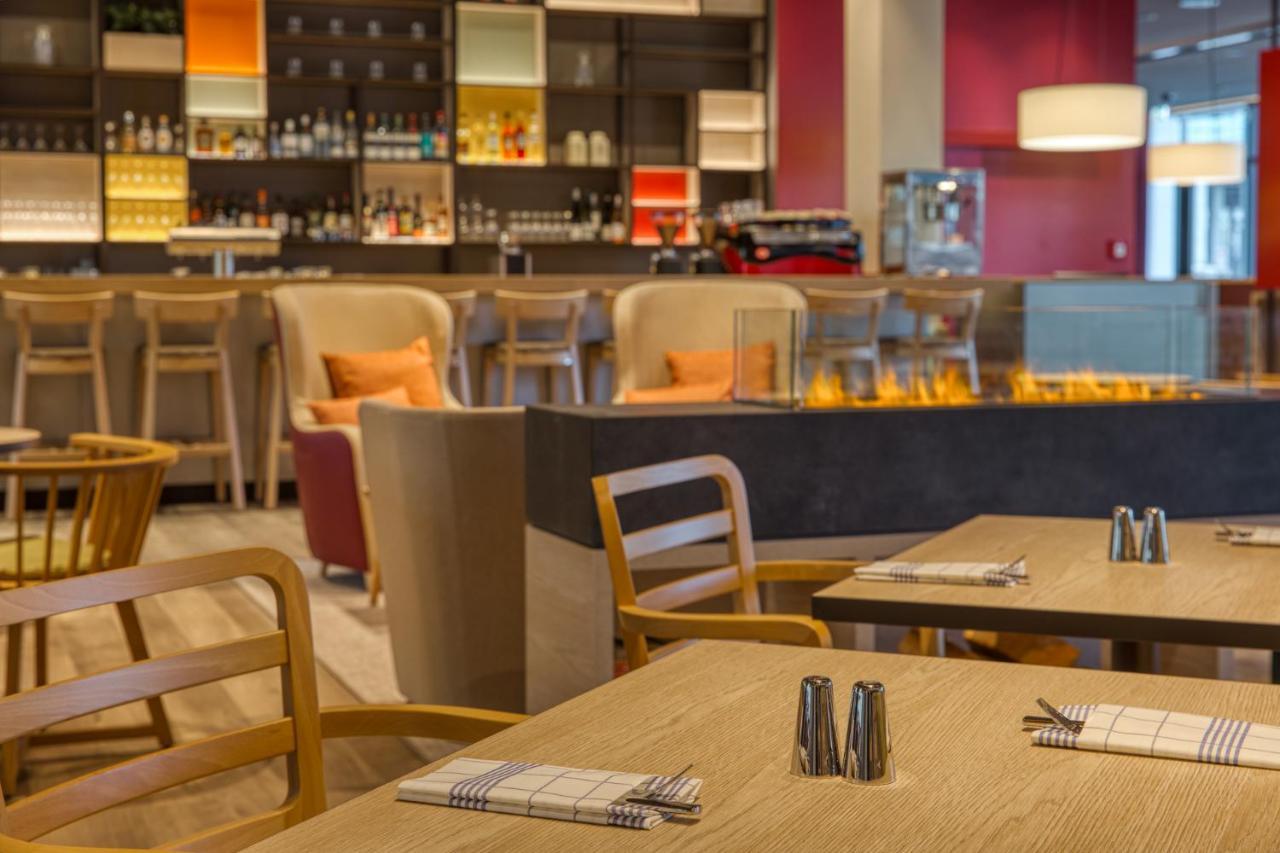 Intercityhotel Berlin Airport Ber Terminal 1 & 2 쇼네펠드 외부 사진 The photo shows a modern restaurant or bar interior. In the foreground, there are wooden tables set with napkins and condiments. The seating includes wooden chairs and plush seating in various colors in the background. A stylish bar area is visible w